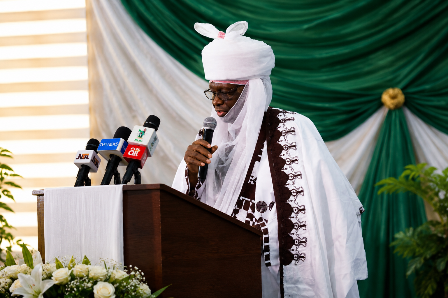 Man in traditional attire speaking at a podium during an event at Al-Muhibbah Open University.